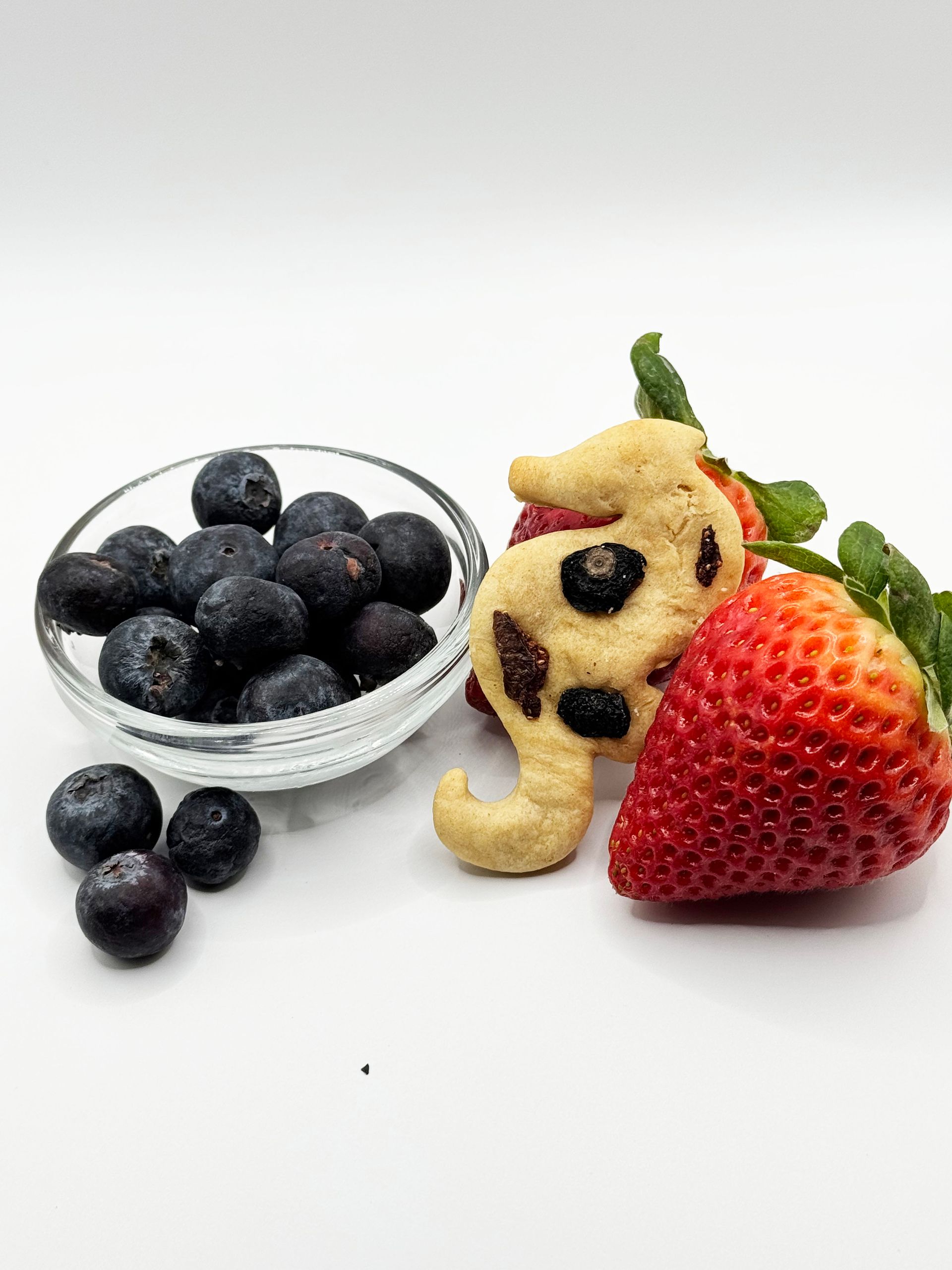 A bowl of blueberries next to a bowl of strawberries and a cookie in the shape of an seahorse