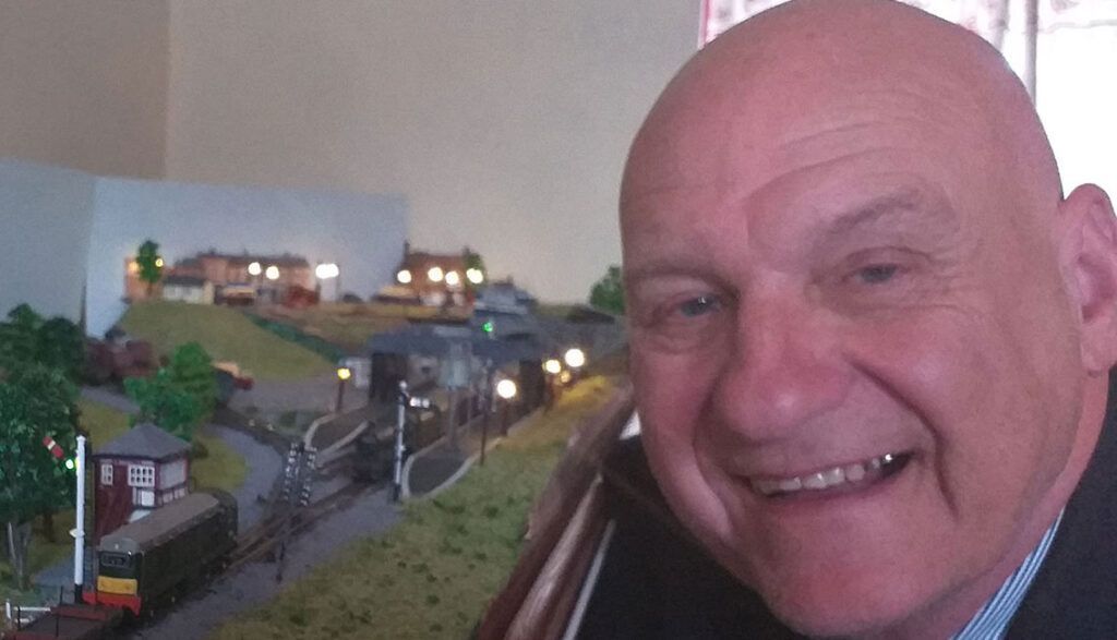 A bald man is smiling in front of a model train track.