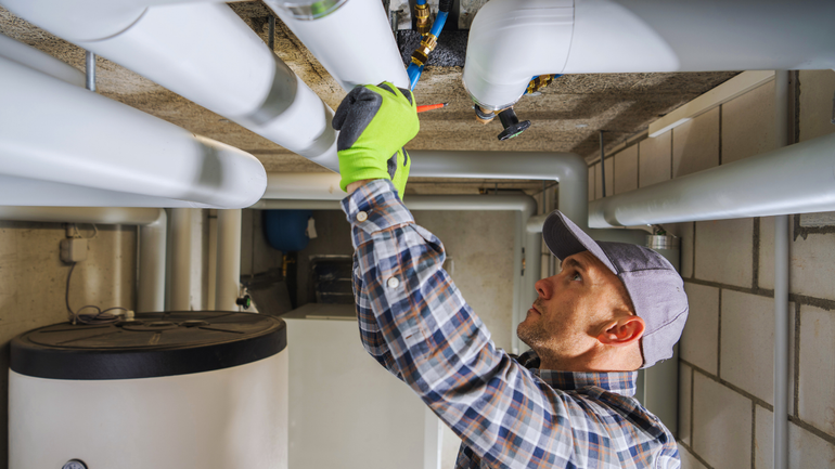 A plumber in a plaid shirt and hat works on white pipes in a basement, wearing gloves.