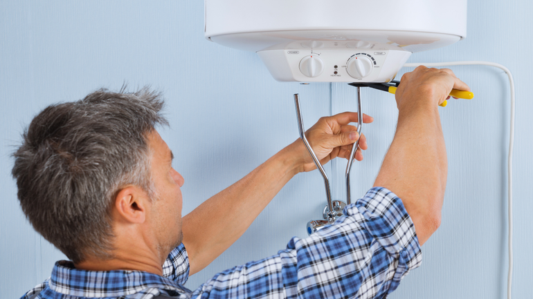 Man in plaid shirt repairs a water heater with pliers, indoors.