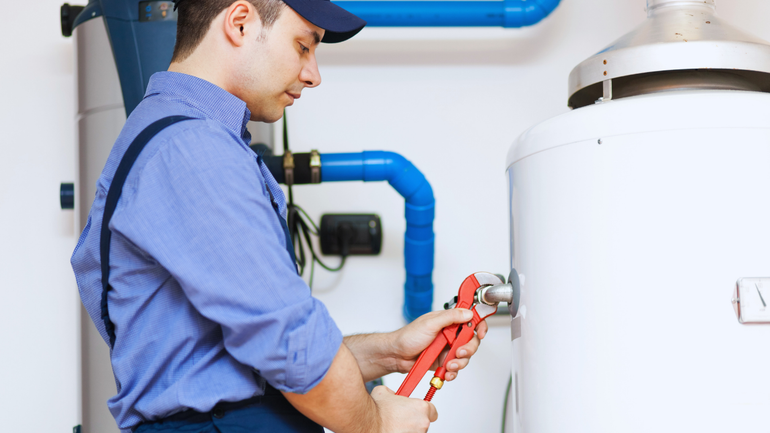 Plumber in blue shirt and overalls, using a wrench on a water heater in a utility room.