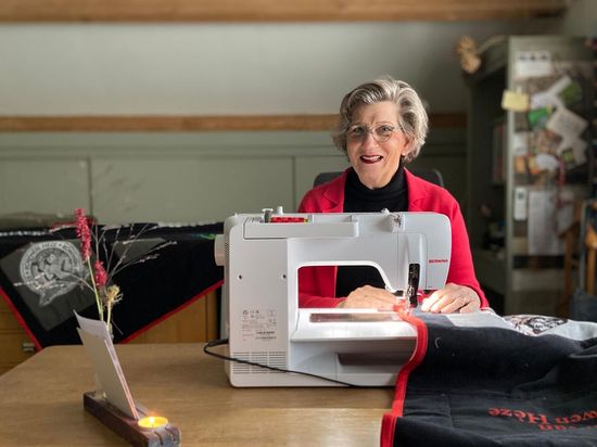 Een persoon, gekleed in een rood vest, zit aan een tafel in een felverlichte kamer en naait stof met een witte naaimachine.