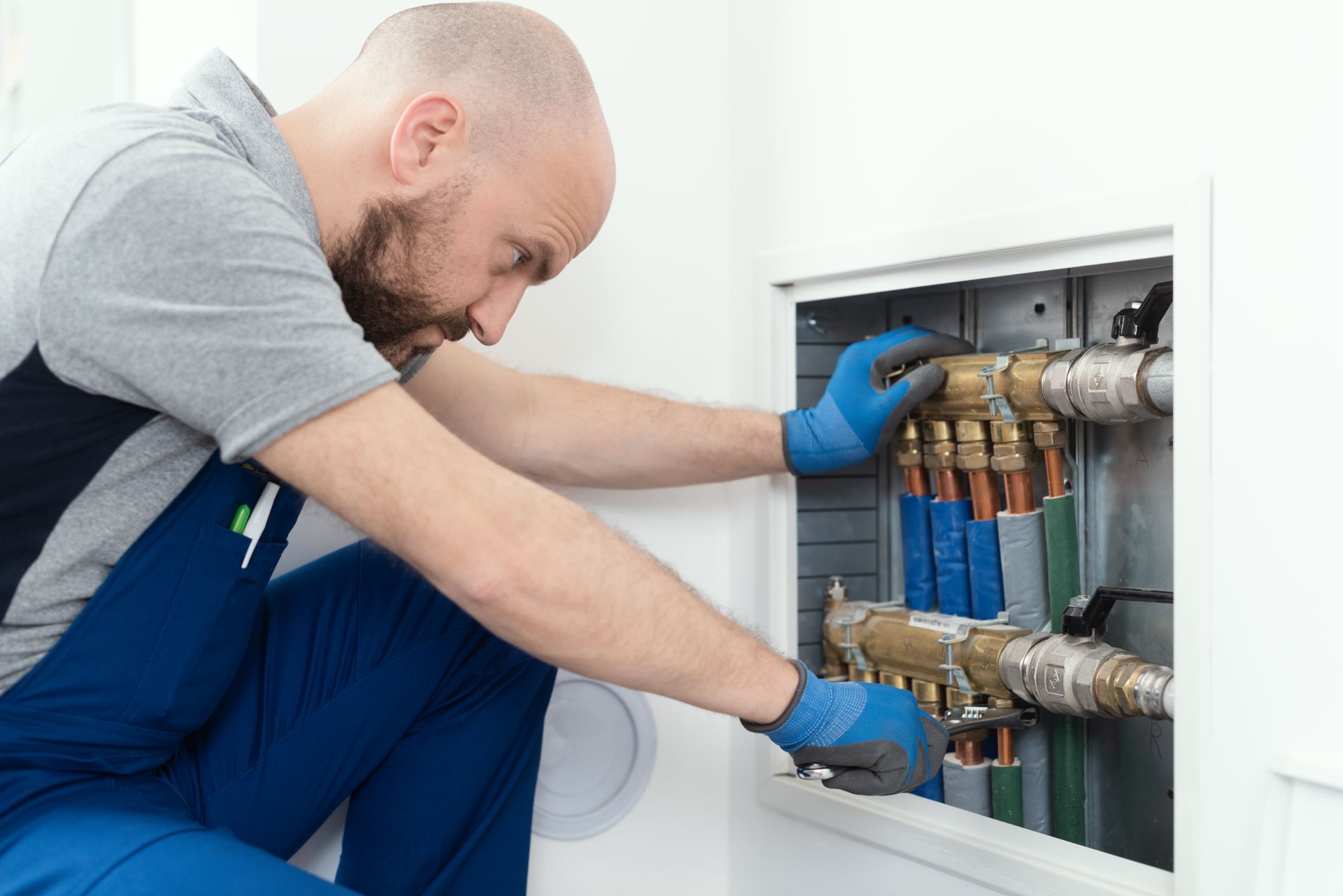 A male residential plumber installs plumbing manifolds while crouching on a tiled white floor.