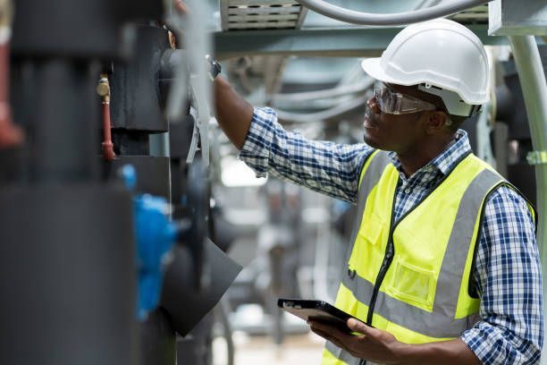 Man in hard hat and safety vest inspecting equipment, holding tablet.