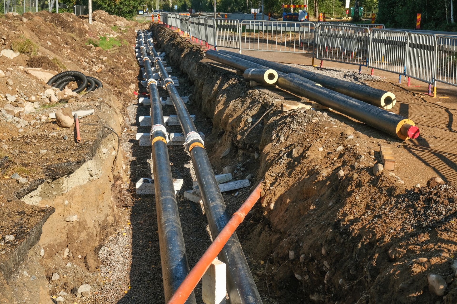 Trench with multiple black pipes being installed, alongside a sidewalk. Earth and support blocks visible.