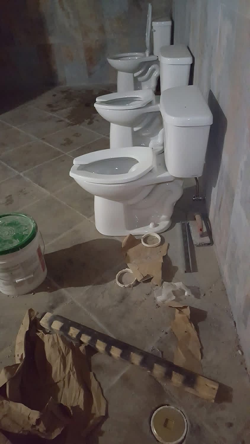 Three white toilets are lined up in a concrete room with scattered debris on the floor.