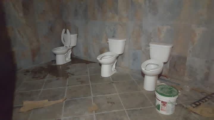 Three toilets in a dimly lit, tiled room with stained walls and floor; a bucket sits near one.