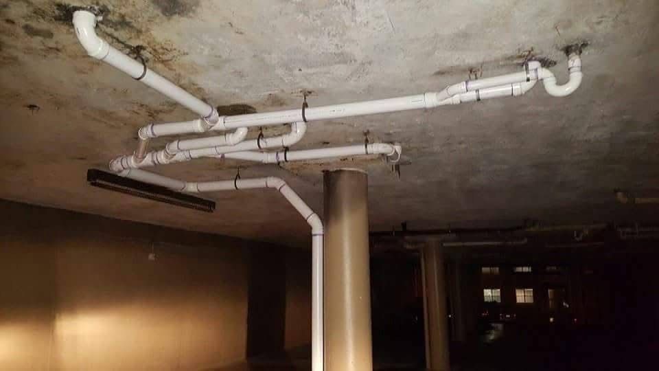 White PVC pipes suspended from a concrete ceiling, likely for plumbing, in a dim basement setting.
