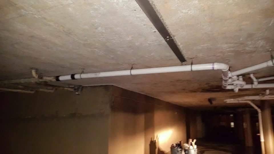 Pipes running across a ceiling in a dimly lit, unfinished space, attached by metal straps.