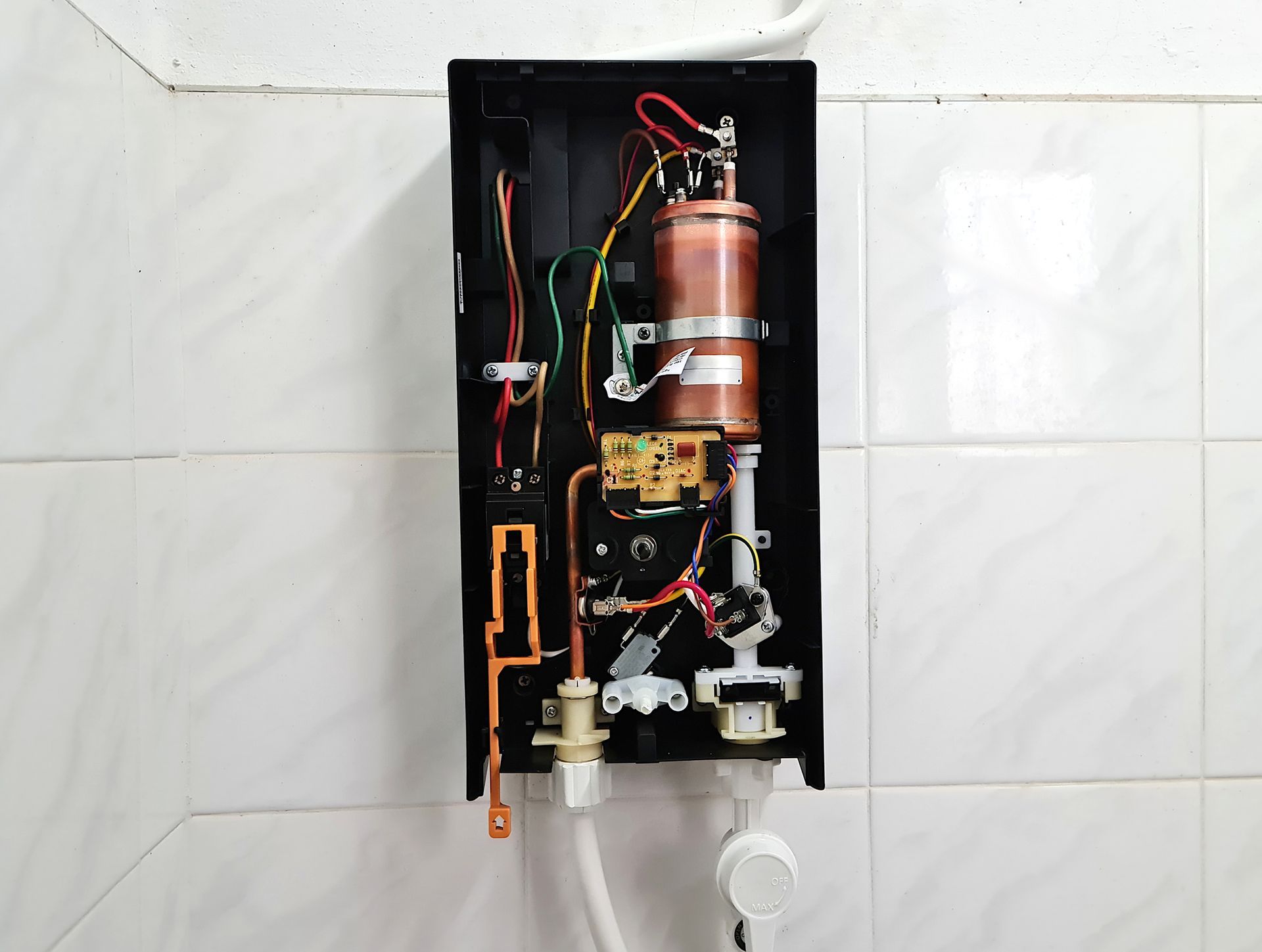 Open electric shower showing internal components on white tiled wall.