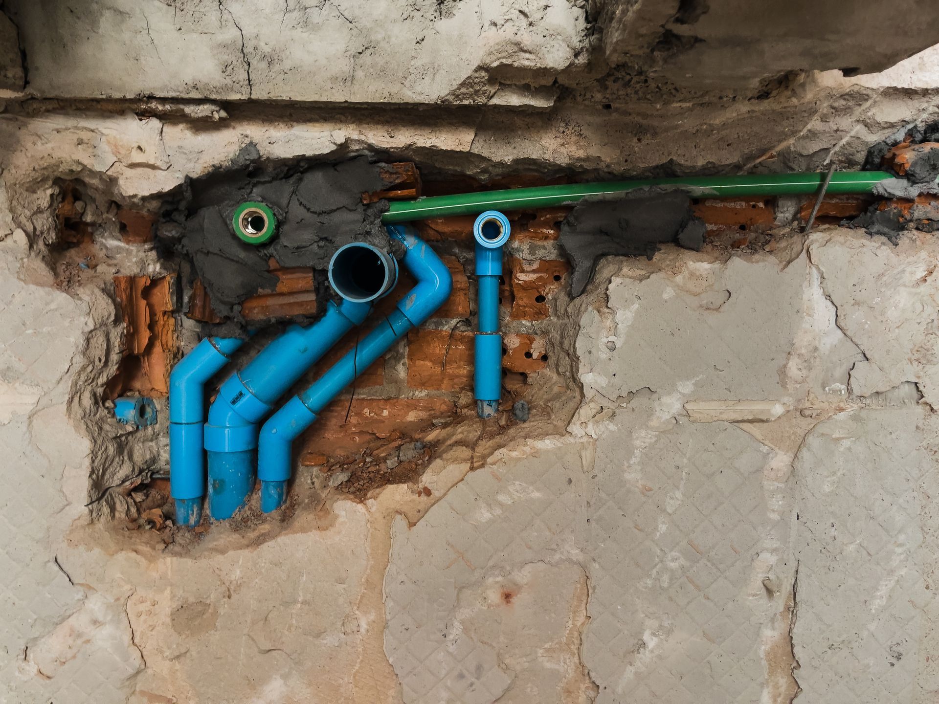 Blue PVC pipes and a green pipe emerge from a damaged brick wall.