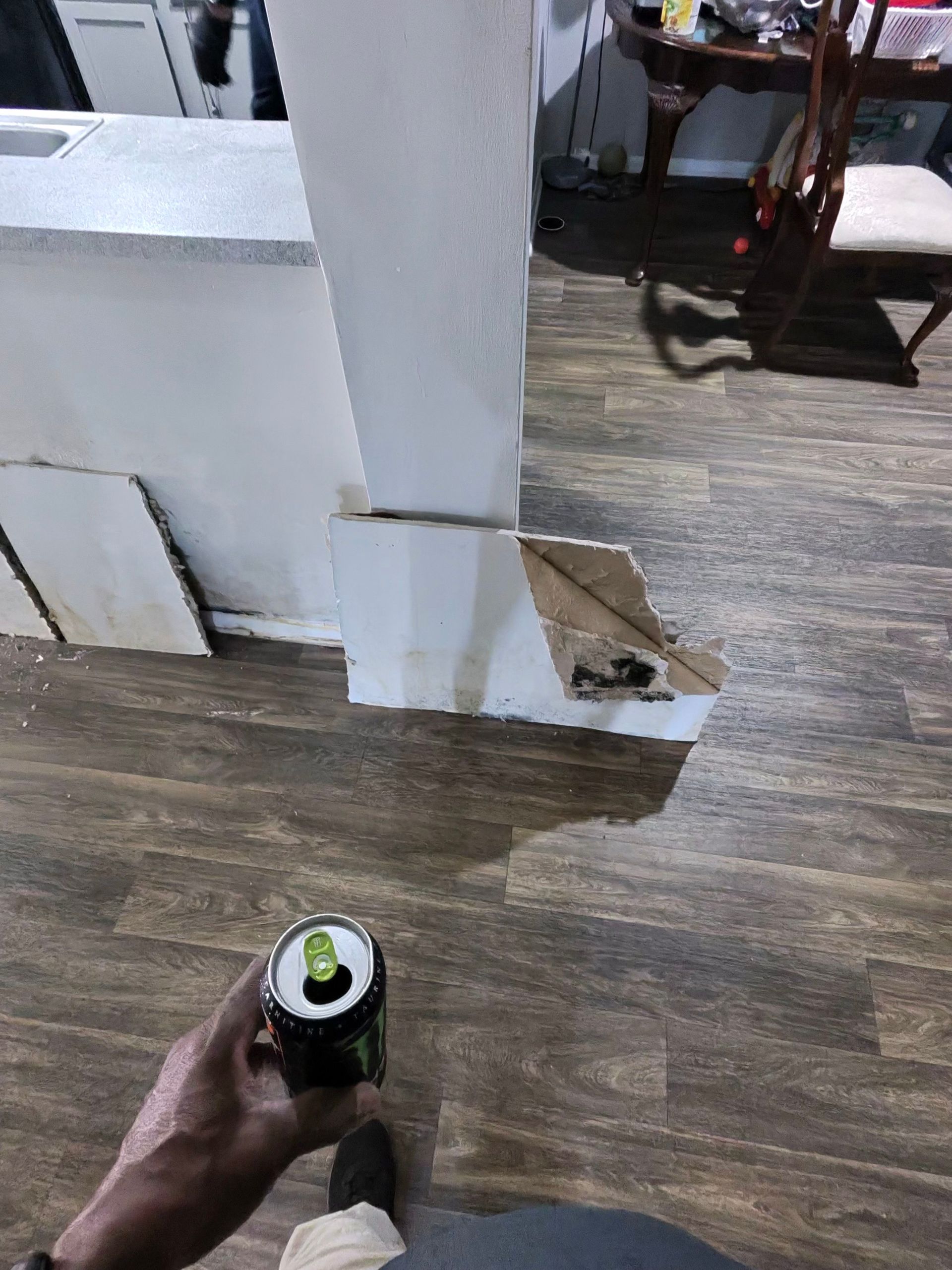 Hand holding an open green and black energy drink can, near a white column and brown floor.