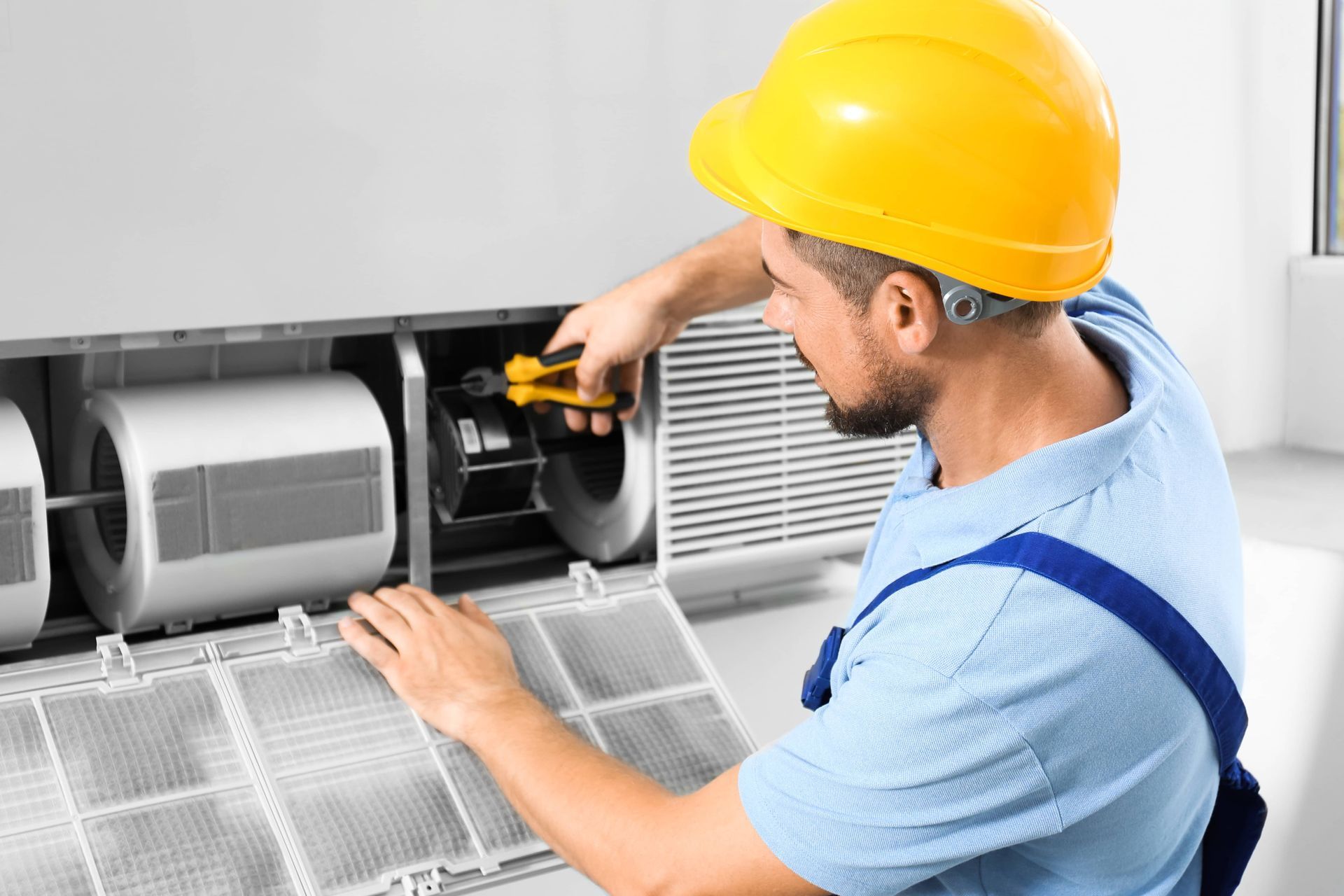 A technician in a yellow hard hat and blue uniform repairs the inner components of a white air conditioning unit.