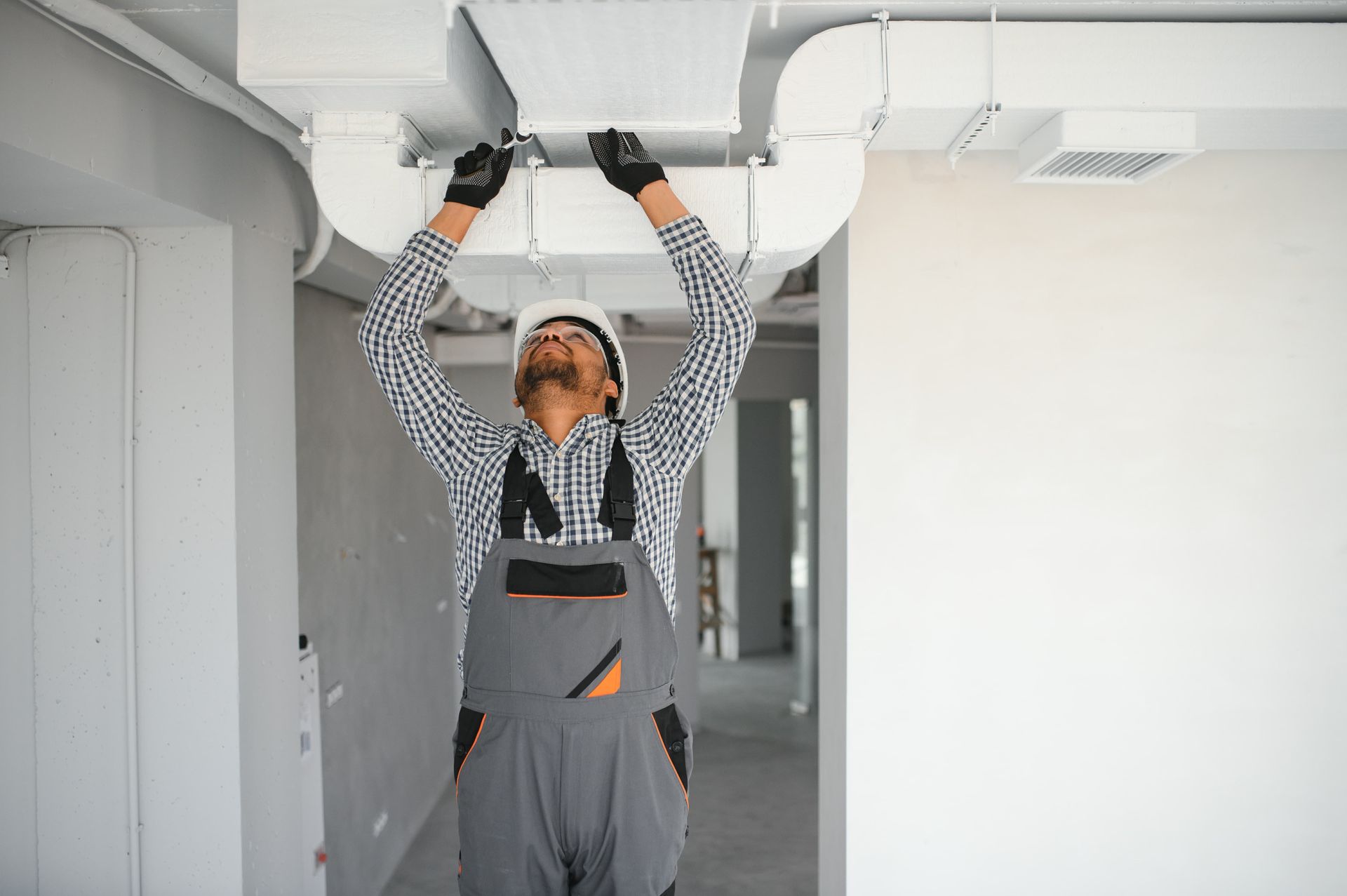 A worker in a white hard hat and grey overalls installs white ventilation ducts on a ceiling in an indoor construction site.