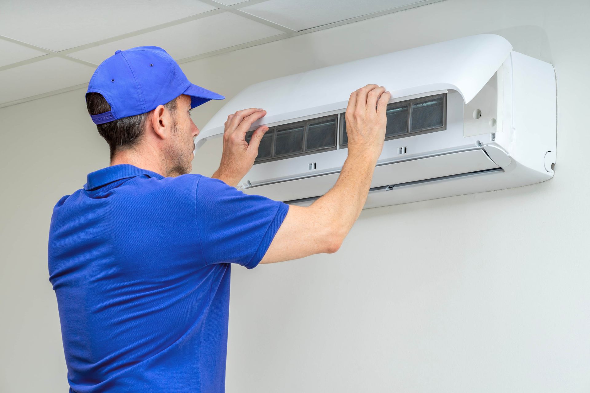A technician in a blue uniform lifting the front panel of a wall-mounted air conditioner to service it.