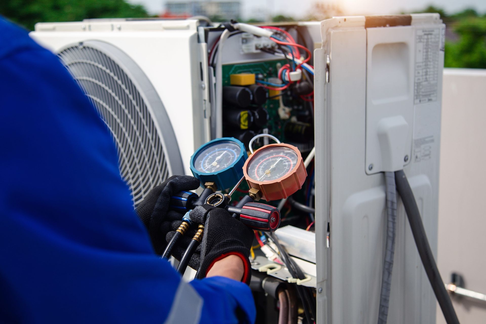 A technician in a blue uniform uses a pressure gauge manifold to service an outdoor air conditioning unit.