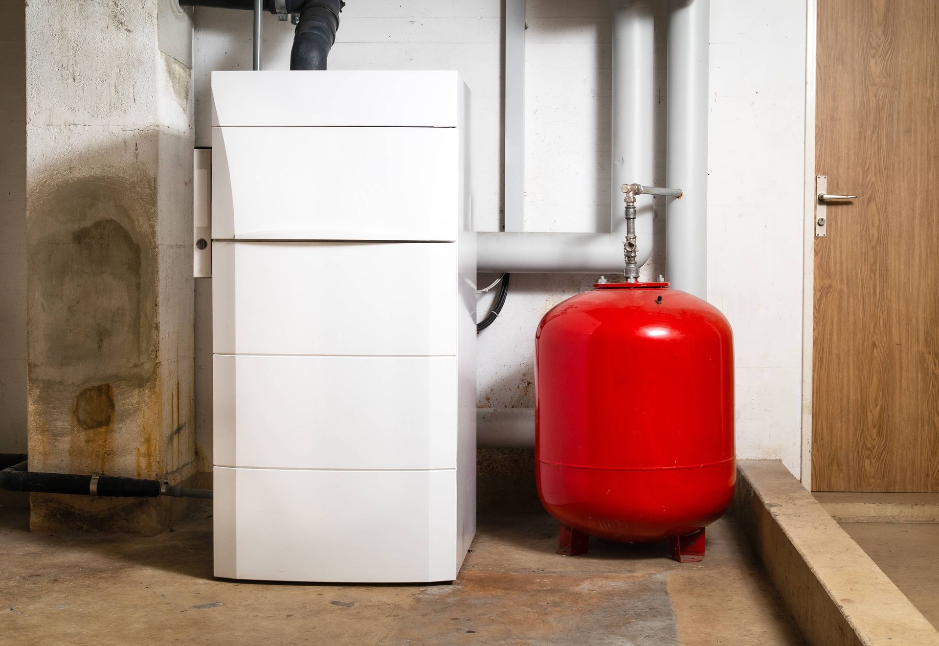 A white heating unit stands next to a red expansion tank in a basement with gray pipes and a wooden door.