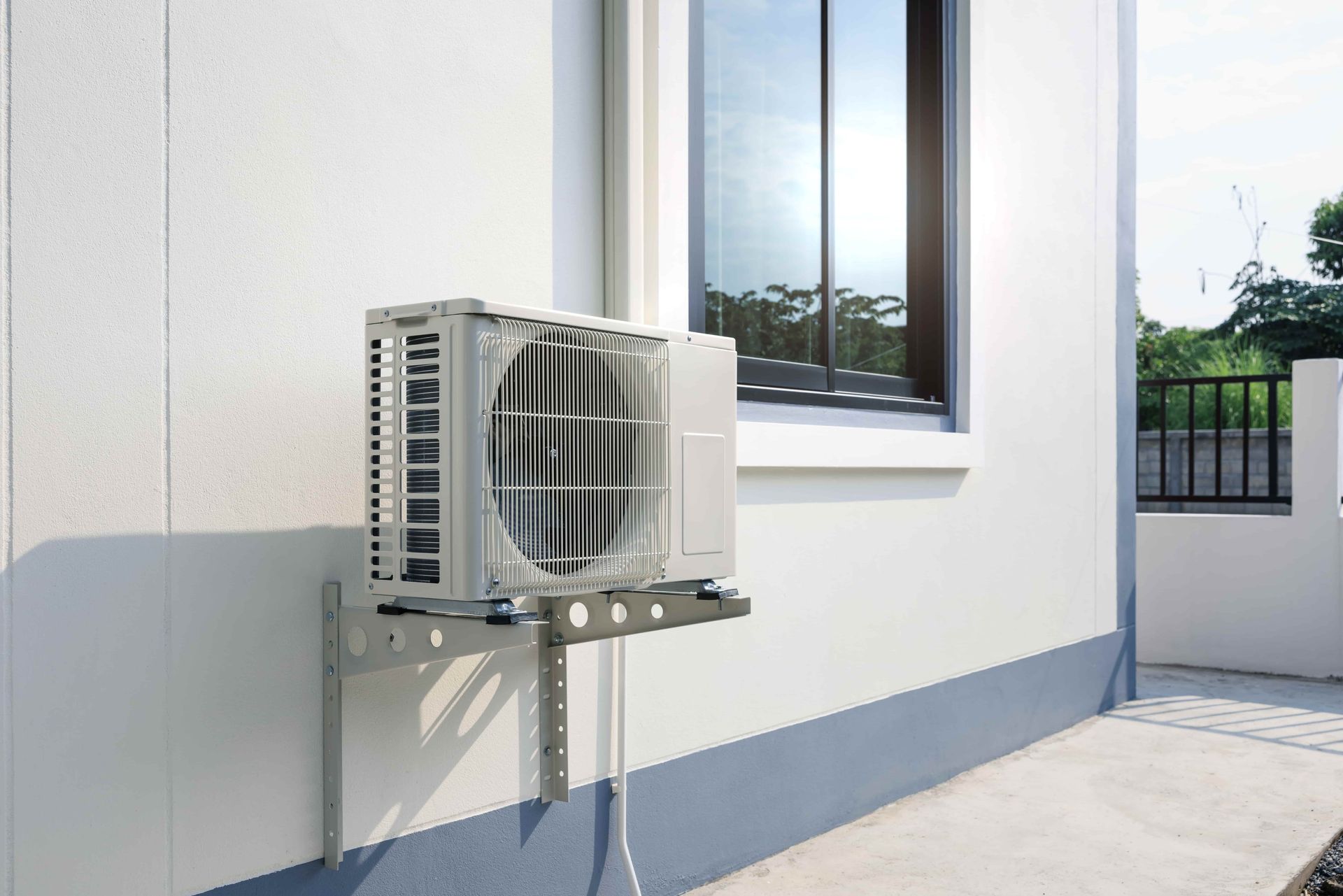 An off-white air conditioner outdoor unit mounted on a wall bracket beside a window on a light-colored building exterior.