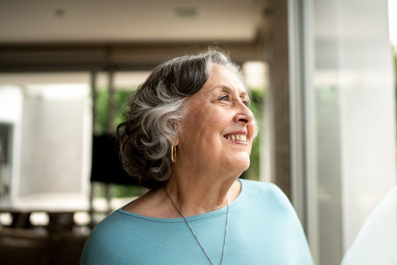 An elderly woman is smiling while looking out of a window.