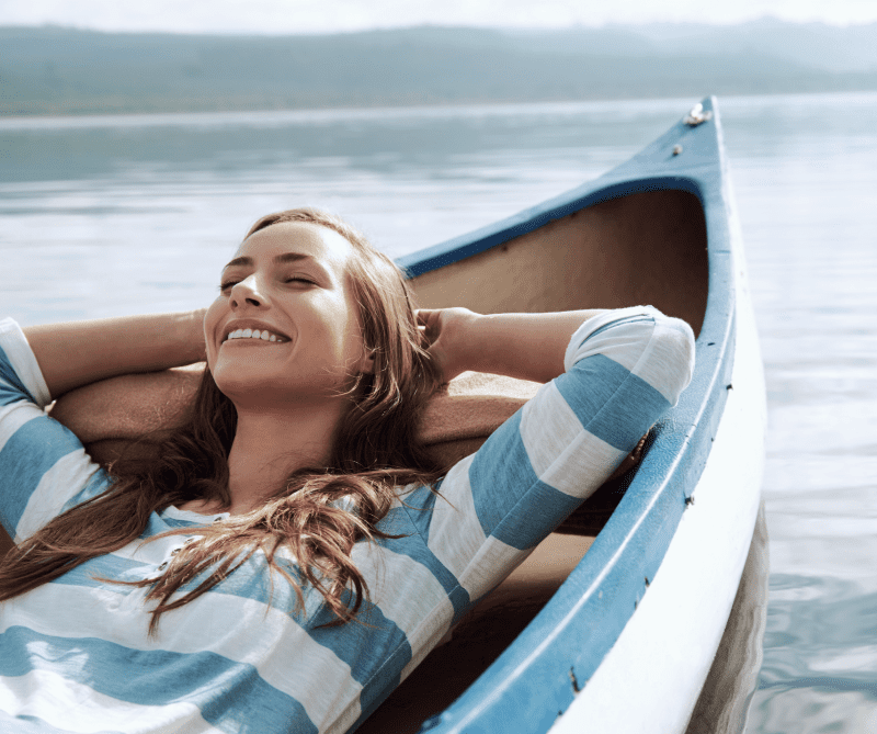 A woman in a striped shirt is laying in a blue canoe on a lake.