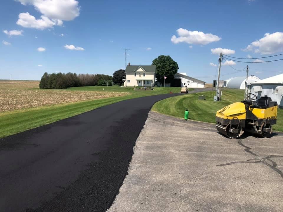 Freshly paved black asphalt driveway leading toward a farmhouse, with a yellow construction roller parked nearby.