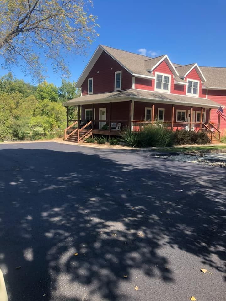 A red two-story house with a wraparound porch and a large, paved driveway under a clear blue sky.