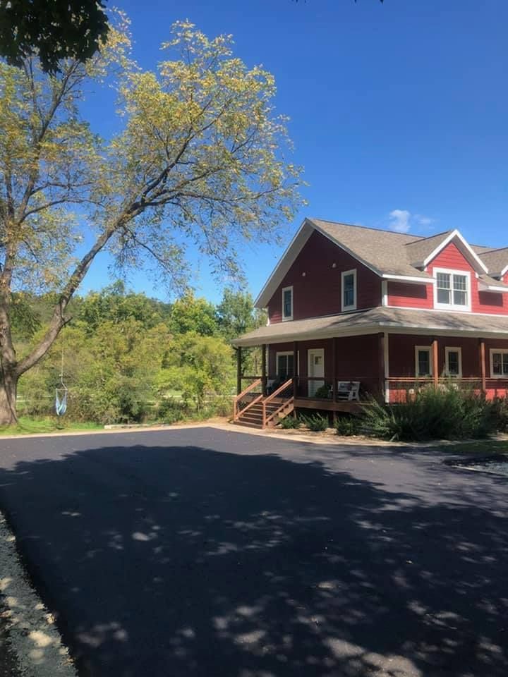 A red house with a wrap-around porch and a large asphalt driveway, set against a backdrop of trees and a blue sky.