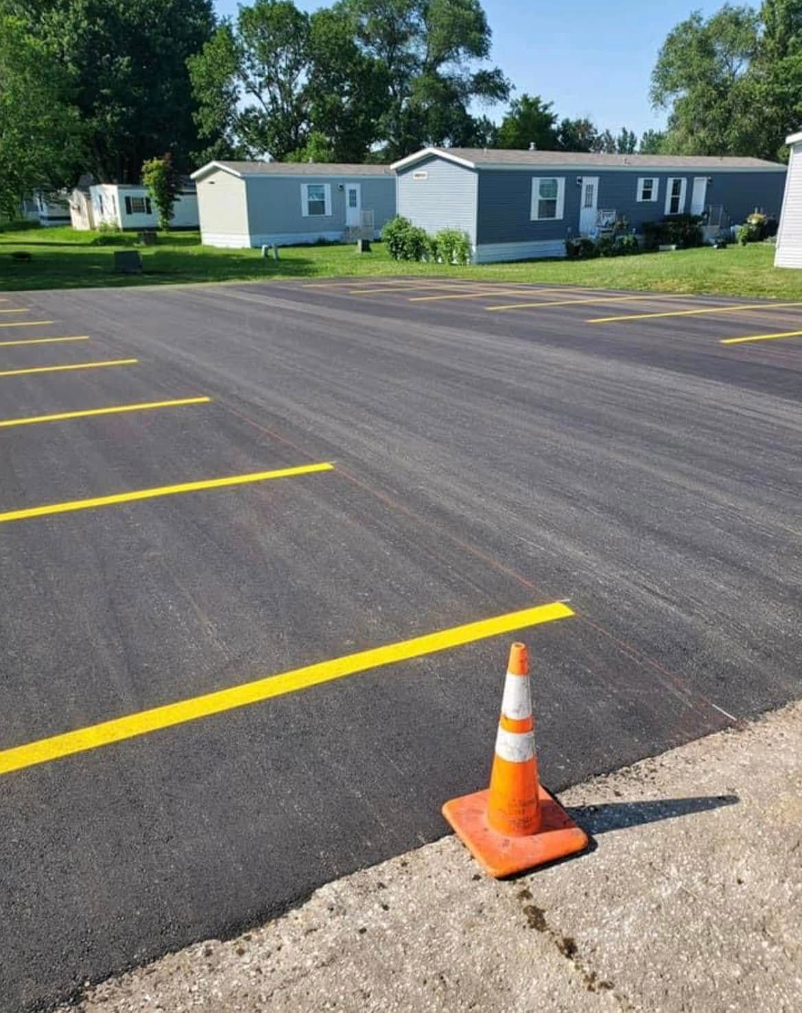 A freshly paved parking lot with painted yellow lines and an orange traffic cone in the foreground, near mobile homes.