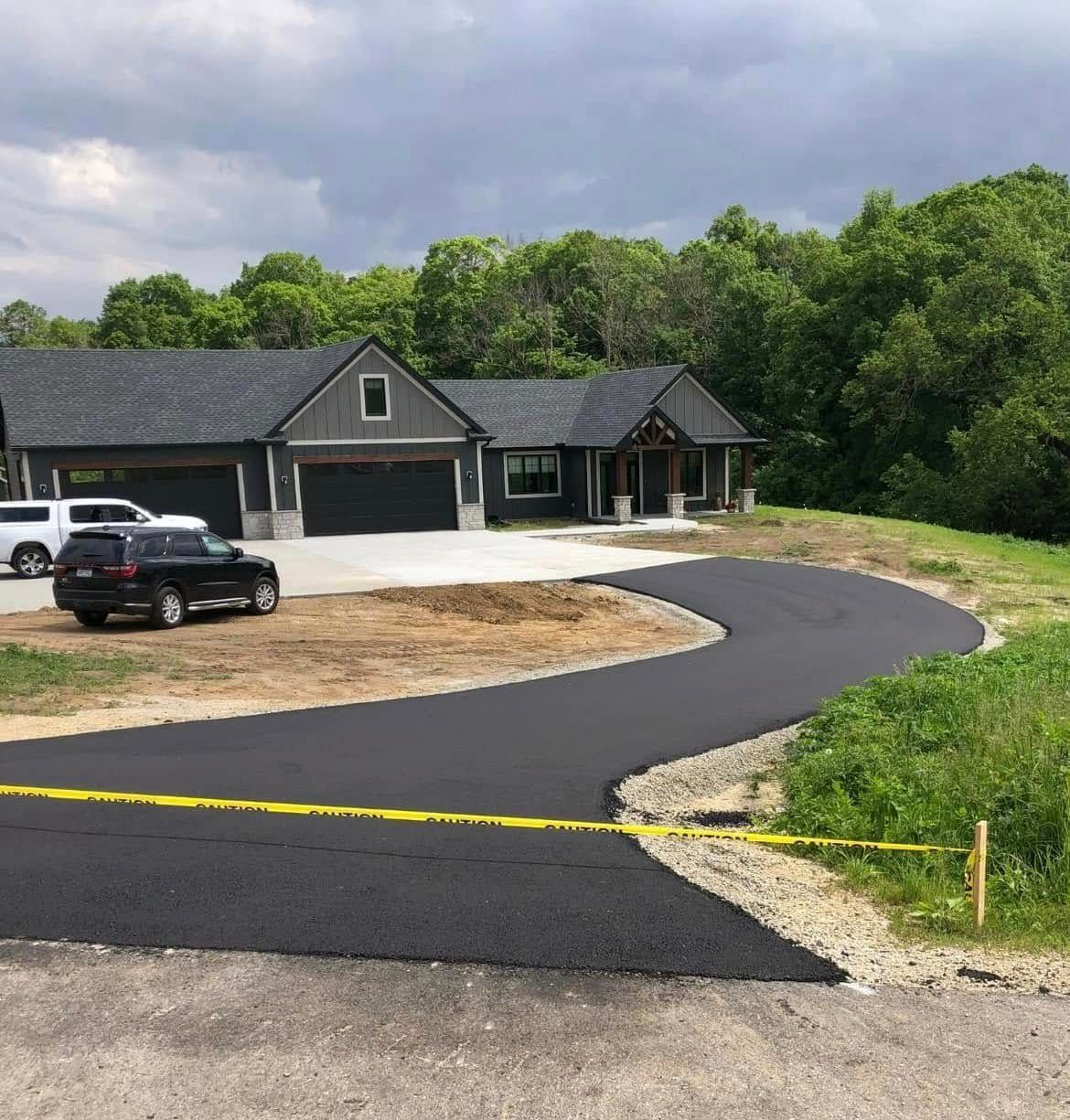 A modern gray house with a three-car garage and a newly paved asphalt driveway in a rural setting with green trees.