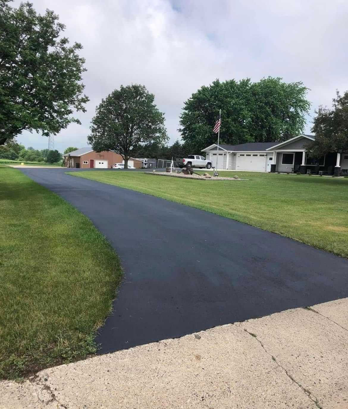 A freshly paved black asphalt driveway leads toward houses on a green lawn under a partly cloudy sky.