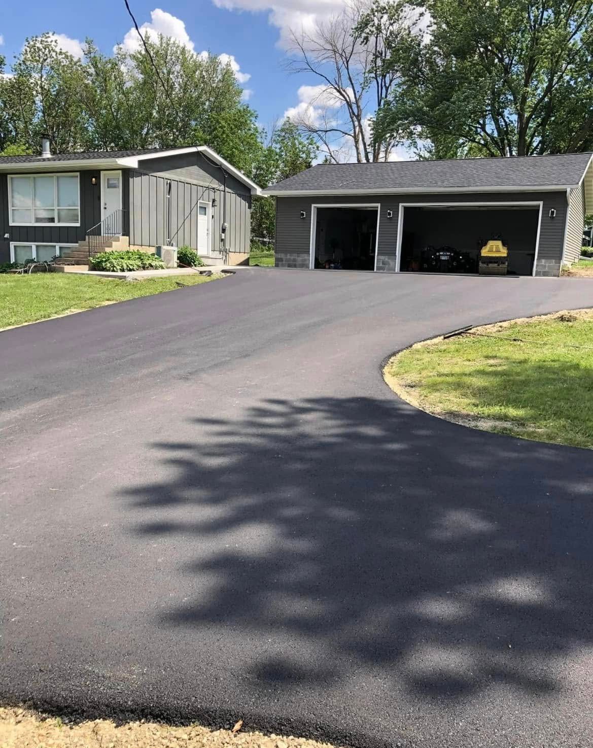 A gray house and detached garage with a freshly paved asphalt driveway under a blue sky with white clouds.