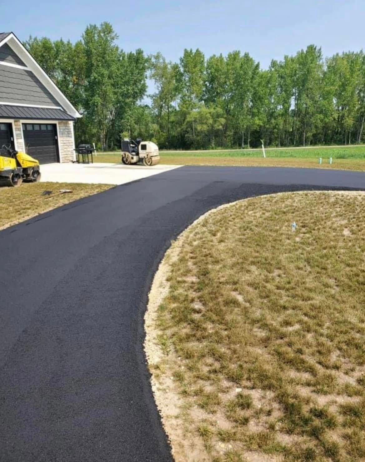 A freshly paved black asphalt driveway curves toward a house with a garage and a construction roller in the background.