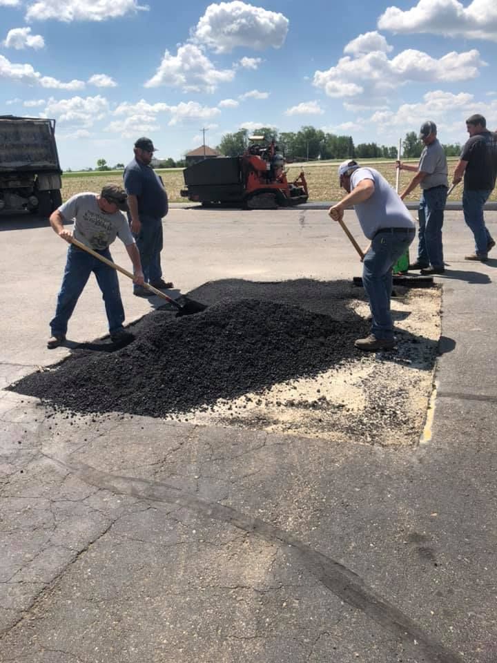 Five workers use shovels and rakes to spread a pile of hot asphalt patch material into a rectangular pothole on a road.