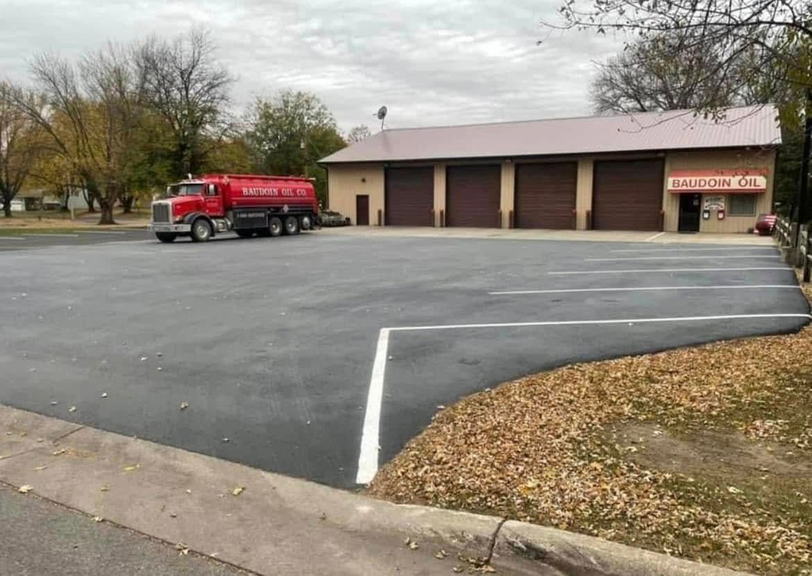 A red tanker truck is parked in a large asphalt lot in front of a tan commercial building with four bay doors.