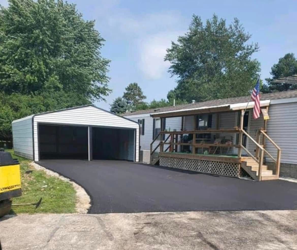 A double-wide carport sits beside a house with a wooden porch and American flag, connected by newly paved black asphalt.