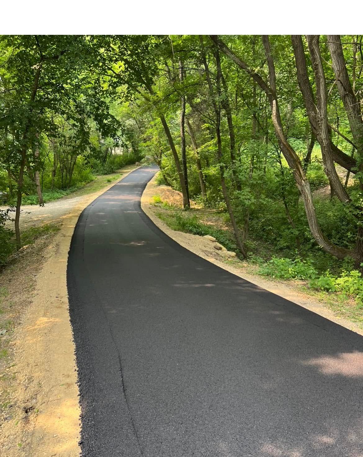 A paved, curving path winds through a lush, green wooded area, flanked by trees and natural dirt shoulders.