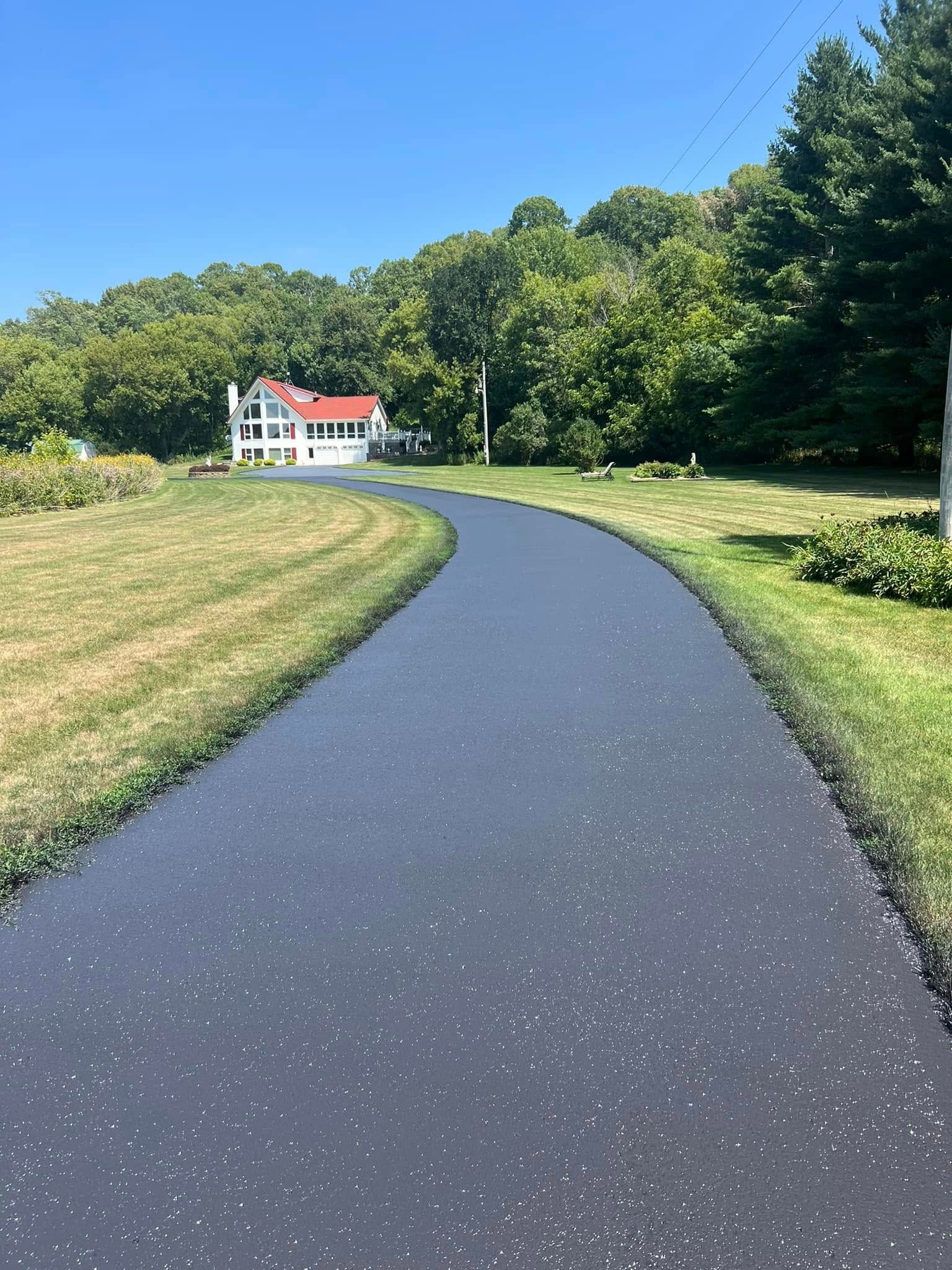 A freshly paved, smooth dark asphalt driveway curving through a green lawn toward a white house with a red roof.