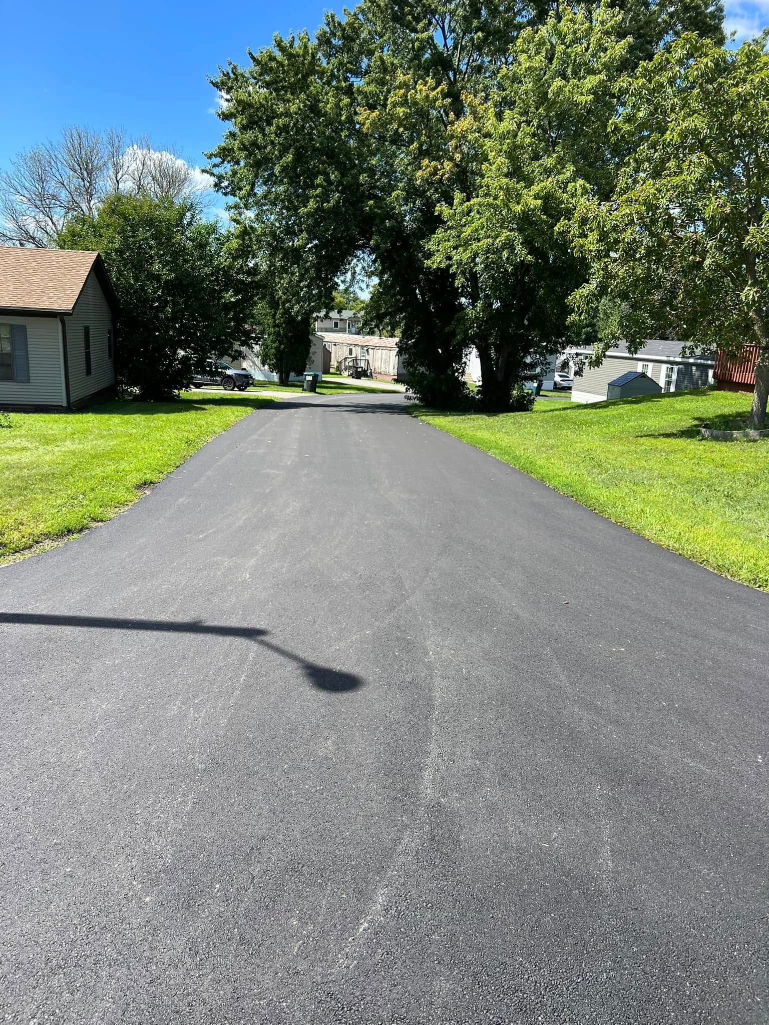 A freshly paved, dark asphalt driveway leads toward a house partially obscured by large, green trees under a blue sky.