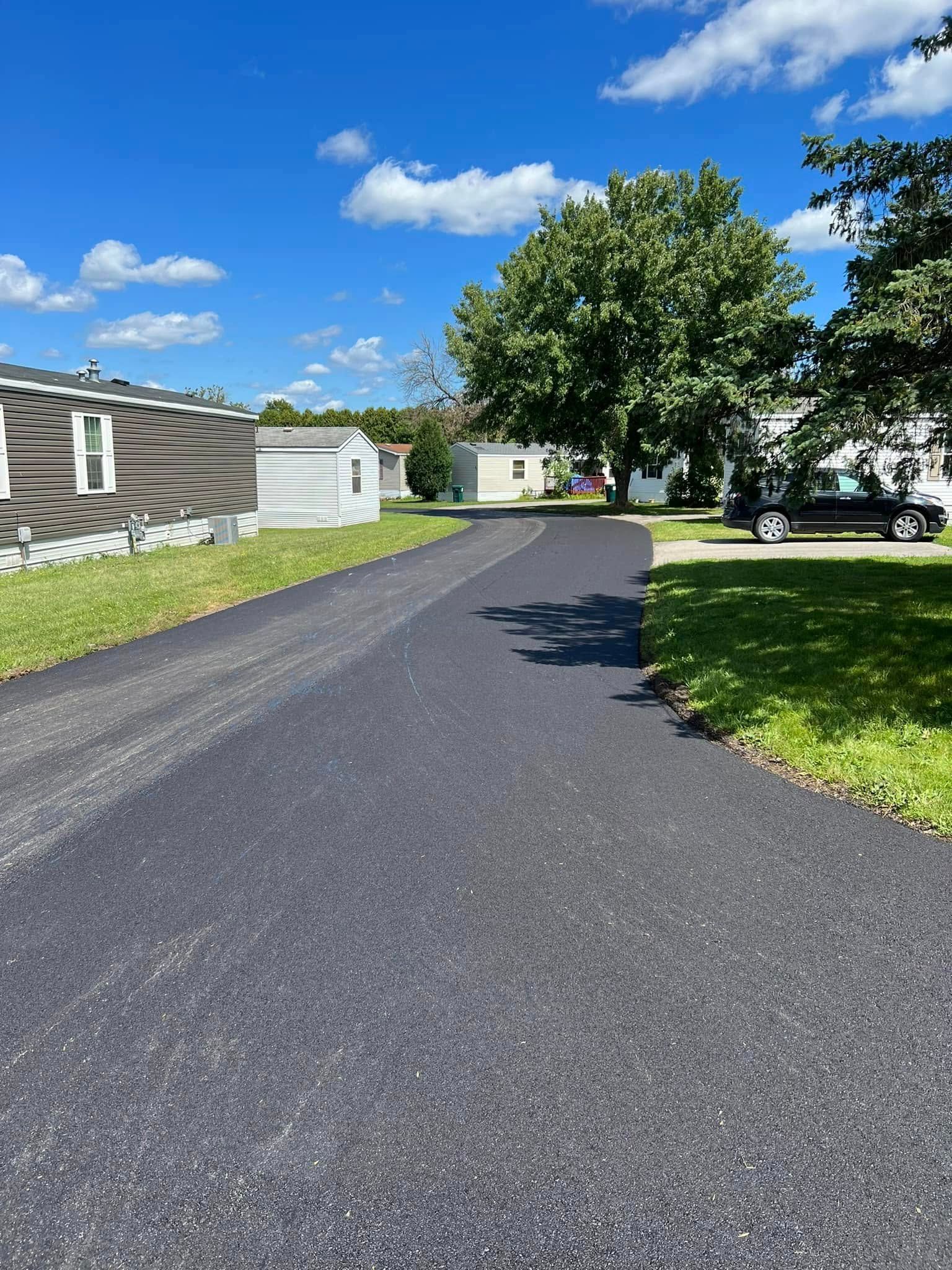 A freshly paved black asphalt driveway curves past mobile homes toward a green tree under a bright blue sky.