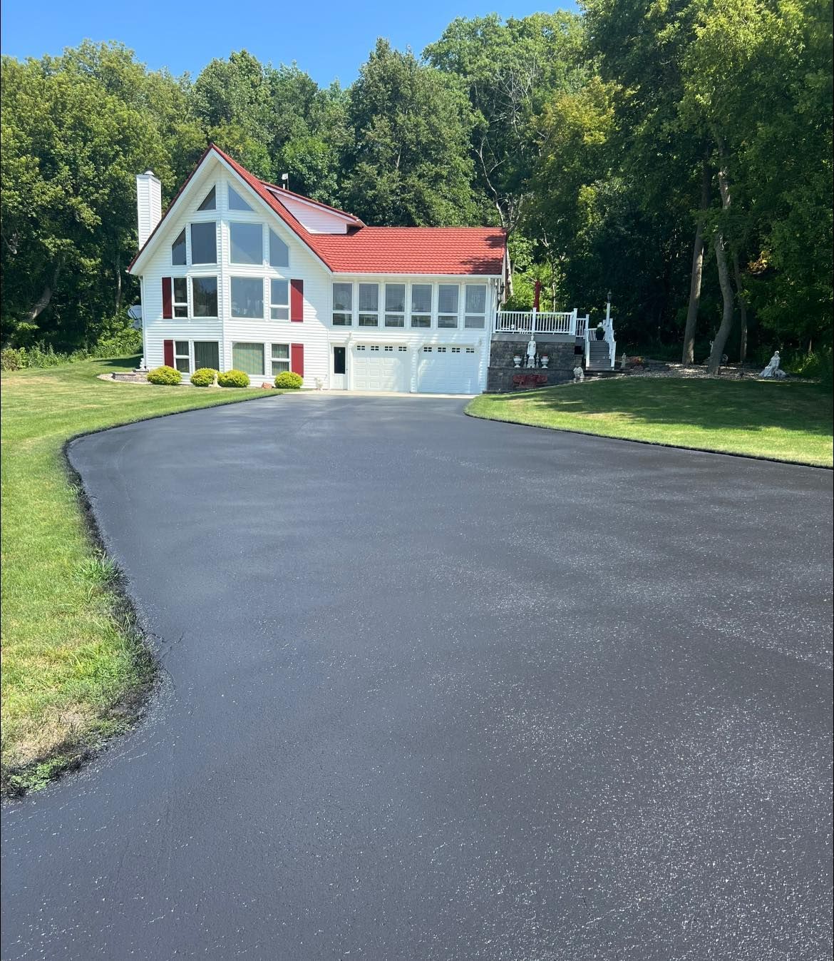 A modern white house with a red roof sits at the end of a freshly paved, smooth black asphalt driveway surrounded by trees.