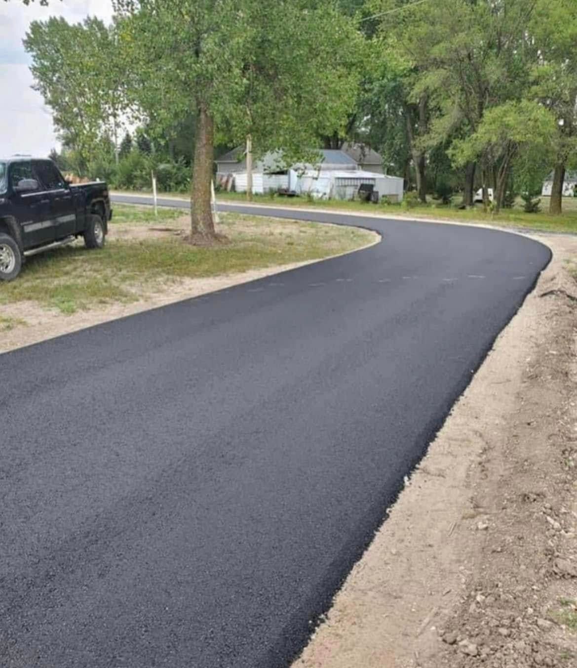A freshly paved black asphalt driveway curves through a residential lawn with trees and a parked truck in the background.