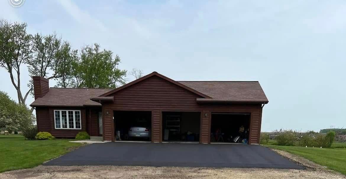 A brown single-story house with a three-car garage and a newly paved asphalt driveway under a clear, light blue sky.