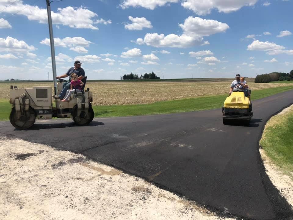 Two people on industrial rollers paving a road through a sunny, open field.