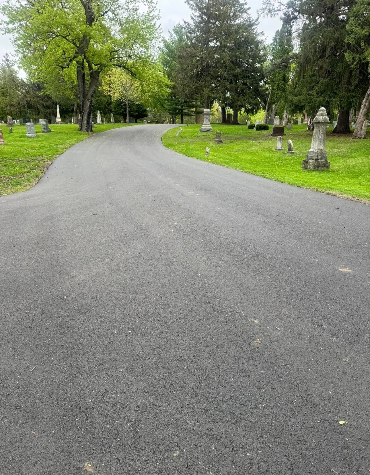 A paved road curves through a cemetery with green grass and trees, featuring various stone markers and monuments.