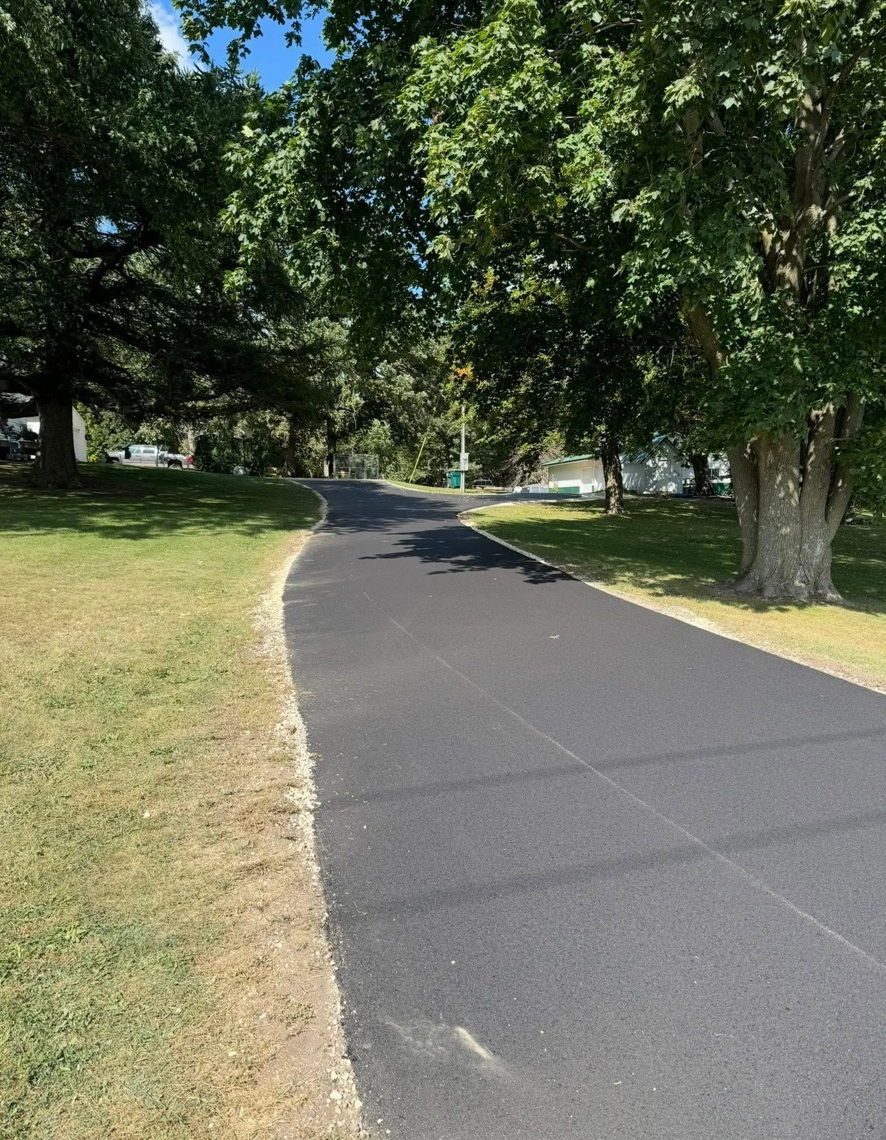 A newly paved, dark asphalt path winds through a grassy park lined with mature green trees under a clear blue sky.