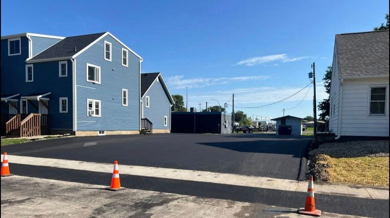A newly paved asphalt driveway sits between a blue multi-story building and a white house under a clear, sunny sky.