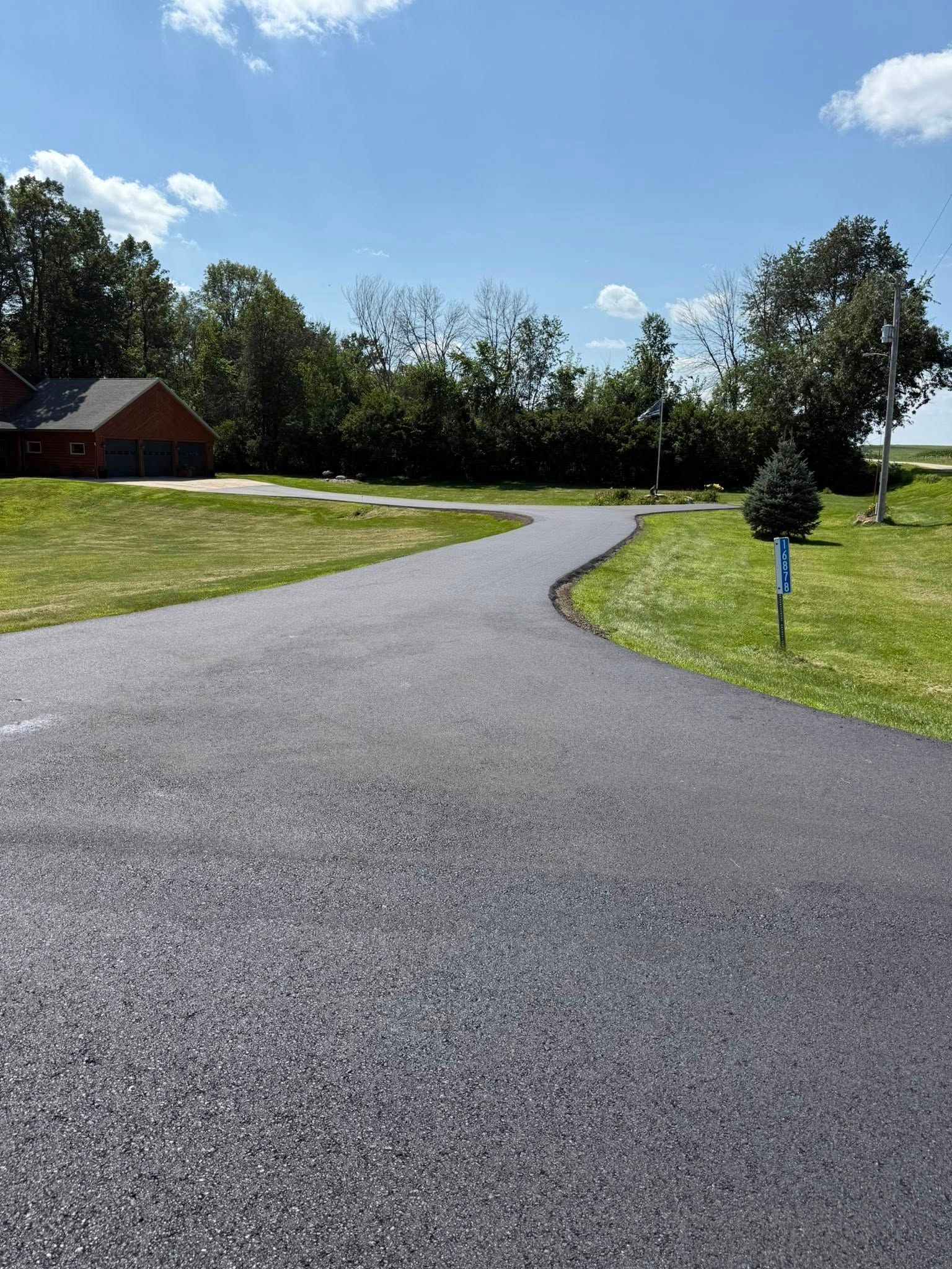 A freshly paved black asphalt driveway curves through a green lawn toward a brick house under a blue, sunny sky.