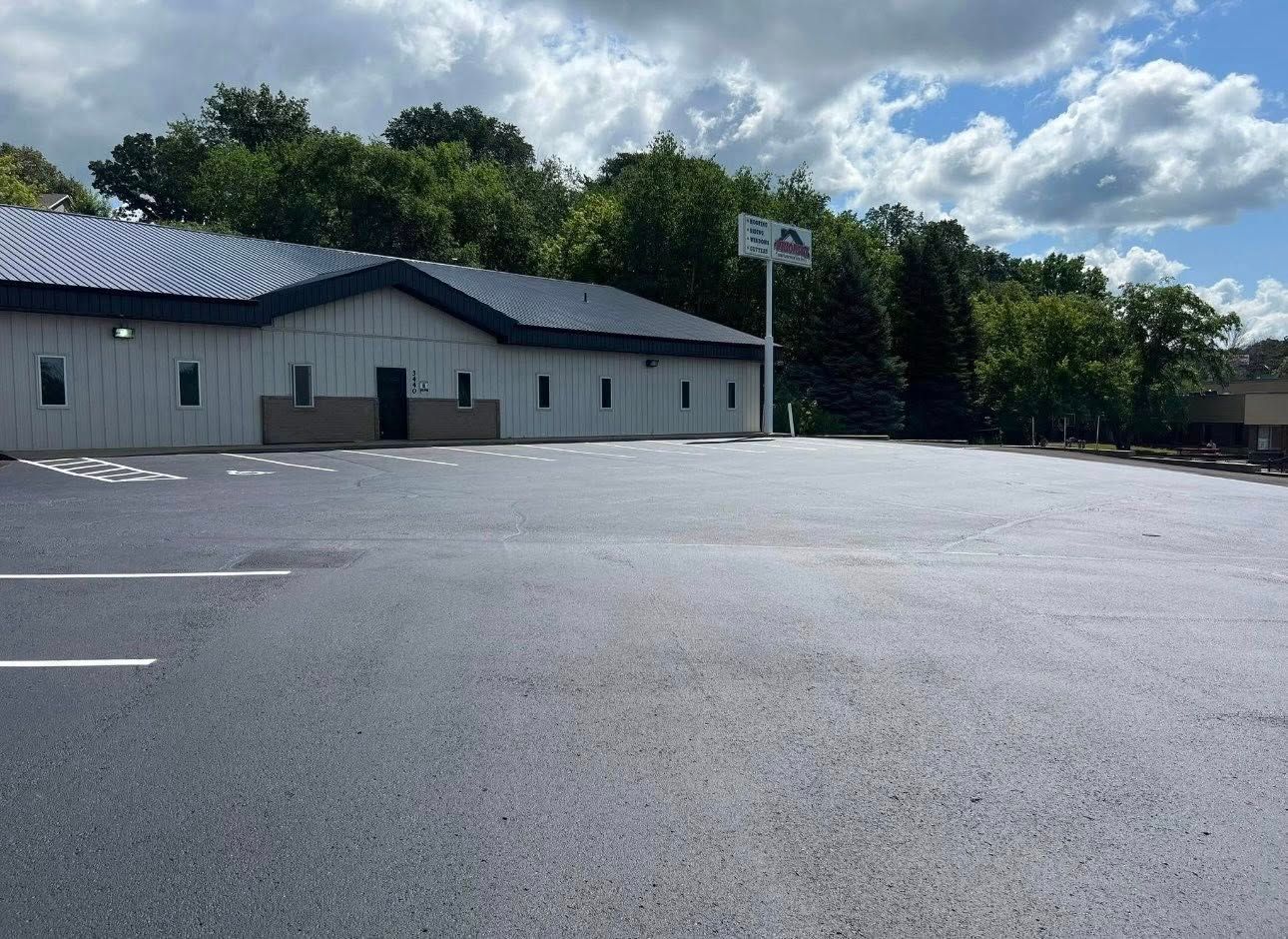 A low, light-colored commercial building with a dark roof stands behind a freshly paved, empty parking lot under a blue sky.