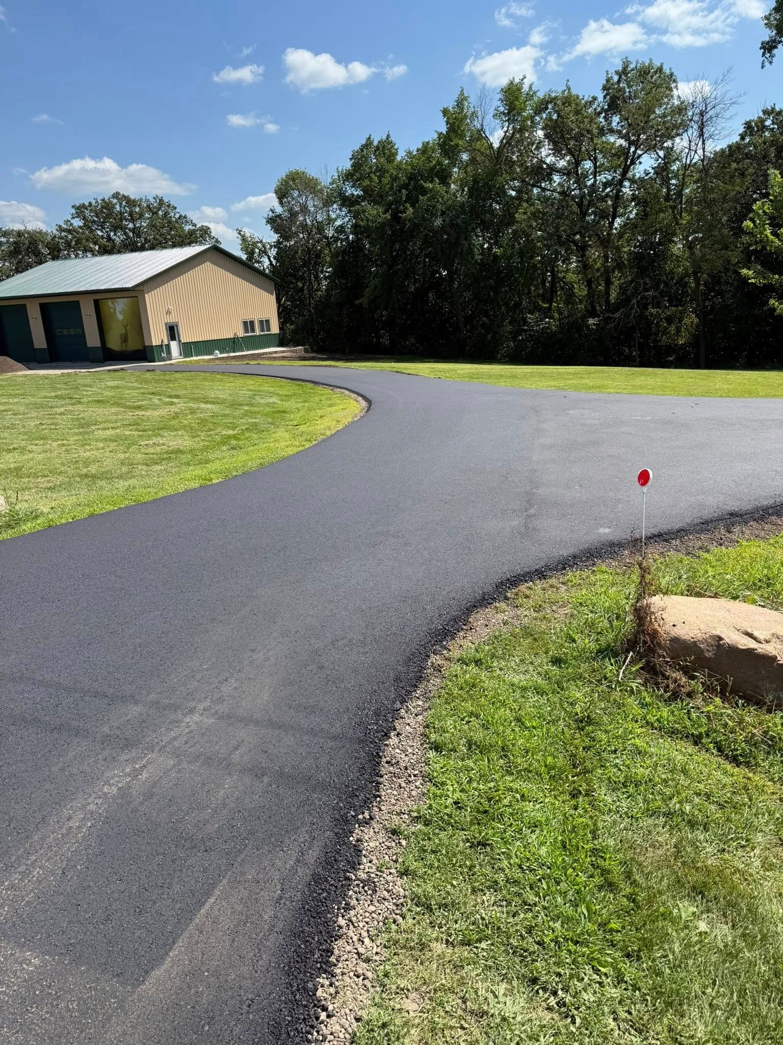 A freshly paved black asphalt driveway curves toward a tan garage building set against a backdrop of green trees.
