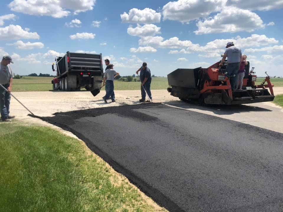 A construction crew uses a paving machine and dump truck to lay fresh black asphalt on a rural road under a sunny sky.